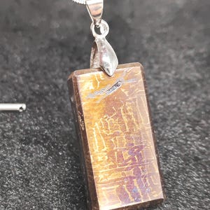 Puede incluir: Un colgante de plata con una piedra rectangular, iridiscente, de color marrón. El colgante tiene una anilla de plata.