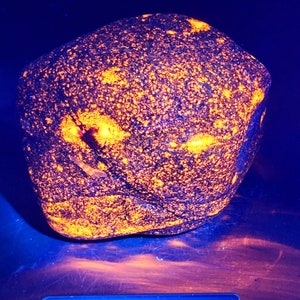 Puede incluir: Una roca azul oscuro con motas y parches de color naranja brillante, iluminada. La roca está sobre una báscula digital, que marca 46,4 kg. La superficie de la roca es texturizada e irregular, con una apariencia única y llamativa.