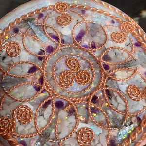 May include: A round, decorative object with a mosaic of white and purple stones, accented with copper wire swirls and floral designs. The edge is trimmed with a twisted copper rope. The center features a copper triskele symbol.