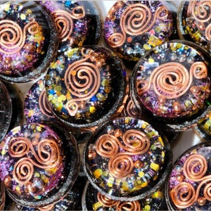 May include: A collection of round, decorative resin pieces. Each piece features a copper wire spiral design, surrounded by colorful glitter and small beads in shades of purple, yellow, and blue. The background is black.