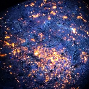 May include: A close-up of a glowing rock. The rock has a dark blue base colour and is covered in small, bright orange and yellow spots. The surface has a textured appearance, with the illuminated spots creating a starry effect.
