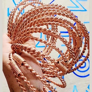 Pode incluir: Uma coleção de pulseiras de metal cor de cobre. As pulseiras têm um design de corda torcida e são empilhadas. O fundo tem formas azuis abstratas. As pulseiras são um acessório único.