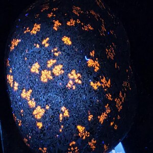 Puede incluir: Una roca oscura y texturizada con patrones vibrantes de color naranja y amarillo, iluminada sobre un fondo negro. Los patrones se asemejan a racimos de flores. La roca está sobre una balanza digital.