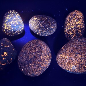 Puede incluir: Seis rocas lisas y redondeadas con un color base azul oscuro. Las rocas están salpicadas de pequeños puntos brillantes de color naranja y amarillo, creando un efecto brillante. Las rocas varían en tamaño y forma, con un fondo azul oscuro.