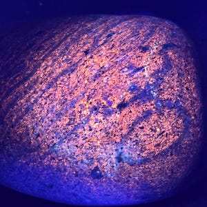 Puede incluir: Primer plano de una roca redondeada con una superficie texturizada. La roca tiene una mezcla de tonos naranja y púrpura, con vetas y parches de color. La iluminación crea un efecto brillante, resaltando los patrones y texturas únicos de la roca.