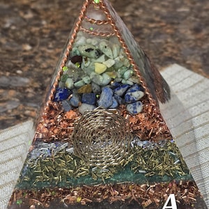 May include: A handcrafted pyramid-shaped decorative object. The pyramid is made of clear resin and features layers of colorful stones, copper shavings, and a gold-colored wire design. The letter "A" is visible on the bottom corner.