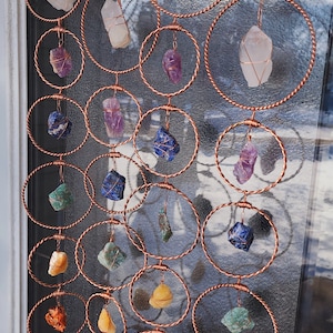 May include: A decorative hanging mobile with raw gemstones, such as quartz, amethyst, and lapis lazuli, suspended within copper wire rings. The crystals are of different shapes and colours, creating a unique and eye-catching display for a window or wall.