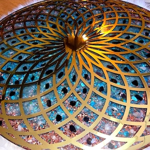 Puede incluir: Un objeto decorativo redondo con una superposición geométrica dorada. El interior presenta un mosaico de piedras azules, marrones y negras. El diseño recuerda a un mandala o un cuenco decorativo, adecuado para la decoración del hogar.
