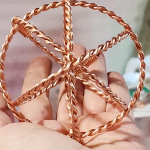 Puede incluir: Una escultura de alambre hecha a mano, de color cobre, con forma de estrella o rueda. El alambre está retorcido, creando una textura similar a una cuerda. La escultura está sostenida en la mano de una persona. El nombre del artista, Jen Lin, está en la esquina inferior derecha.