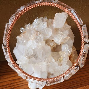 Puede incluir: Un círculo de alambre de cobre adornado con cristales de cuarzo transparente, que rodea un gran grupo de cristales de cuarzo natural. Los cristales son transparentes y tienen una textura natural y rugosa. El alambre de cobre está bien enrollado.