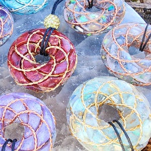 Può includere: Primo piano di diversi ciondoli a forma di ciambella in vari colori, tra cui rosso, blu e viola. Ogni ciondolo presenta un design in filo di rame intrecciato e un cordino nero. Alcuni ciondoli hanno un aspetto glitterato e iridescente, mentre altri sono più uniformi nel colore. I ciondoli sono esposti su una superficie chiara.