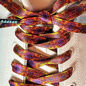 May include: Close-up of white sneakers with vibrant, patterned shoelaces. The laces feature a repeating floral design in shades of orange, yellow, and purple. A small metal tag with the word "SPIFFY" is visible on the laces.