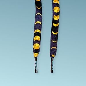Puede incluir: Dos cordones de zapatos negros con un patrón repetido de fases lunares amarillas y doradas sobre un fondo azul oscuro. Las puntas de los cordones son negras con la palabra "SPIFFY" impresa en azul claro.