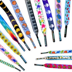 May include: A collection of flat shoelaces featuring various colorful patterns including moon phases, cookies, piano keys, floral prints, alien faces, donuts, cherries, sunflowers, dinosaurs, pineapples, and frogs, each with a black aglet labeled SPIFFY.