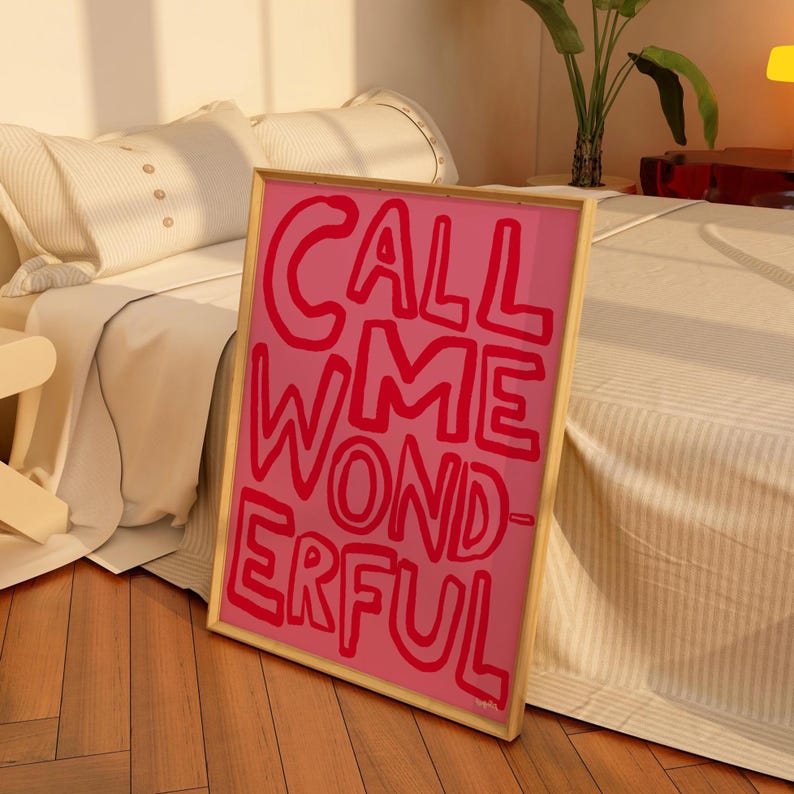 May include: A framed pink art print with the words "CALL ME WONDERFUL" in red, bold, handwritten-style letters. The print has a gold frame and is leaning against a bed with a neutral-colored comforter and pillows.