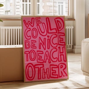以下が含まれることがあります： 「WE COULD BE NICE TO EACH OTHER」という赤い太字の手書き文字が書かれた、額入りのピンク色のアートプリント。アートワークは、壁に立てかけられた明るい色の木製フレームに表示されています。