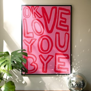 K&ouml;nnte beinhalten: Ein gerahmter rosa Kunstdruck mit den Worten "OK LOVE YOU BYE" in roten Blockbuchstaben. Das Kunstwerk wird &uuml;ber einer Holzoberfl&auml;che mit einer Discokugel und einer gr&uuml;nen Pflanze im Vordergrund angezeigt.