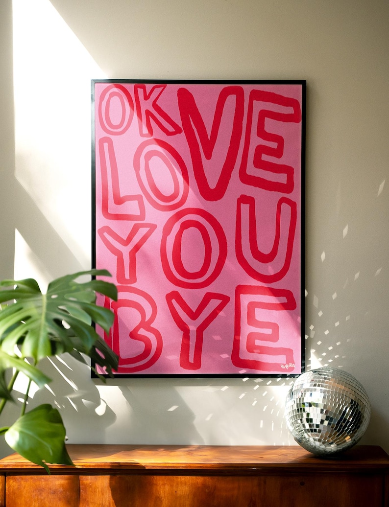 K&ouml;nnte beinhalten: Ein gerahmter rosa Kunstdruck mit den Worten "OK LOVE YOU BYE" in roten Blockbuchstaben. Das Kunstwerk wird &uuml;ber einer Holzoberfl&auml;che mit einer Discokugel und einer gr&uuml;nen Pflanze im Vordergrund angezeigt.