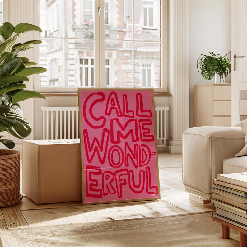 May include: A framed pink poster with the words "CALL ME WONDERFUL" in bold, red letters. The poster is propped up against a cardboard box on a wooden floor. The room has a large window and a neutral color scheme.