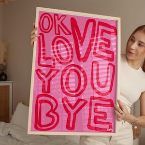 K&ouml;nnte beinhalten: Ein gerahmter rosa Kunstdruck mit den Worten "OK LOVE YOU BYE" in roten, fetten Buchstaben. Das Kunstwerk wird in einem hellfarbigen Holzrahmen pr&auml;sentiert. Der Hintergrund ist einfarbig rosa.