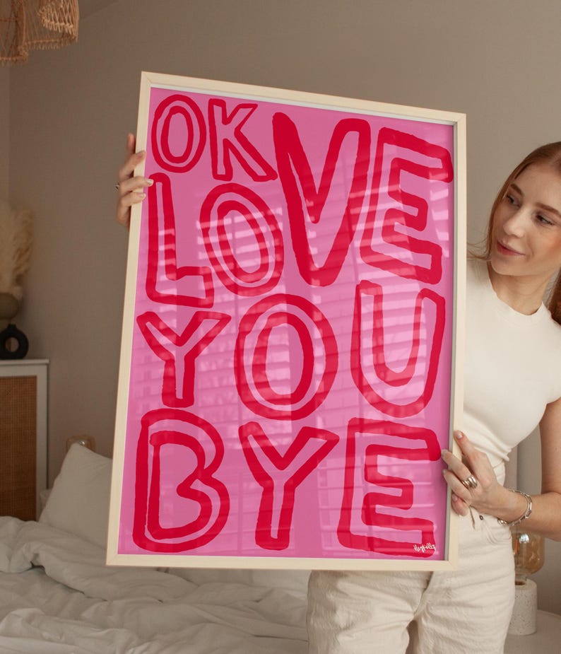 K&ouml;nnte beinhalten: Ein gerahmter rosa Kunstdruck mit den Worten "OK LOVE YOU BYE" in roten, fetten Buchstaben. Das Kunstwerk wird in einem hellfarbigen Holzrahmen pr&auml;sentiert. Der Hintergrund ist einfarbig rosa.