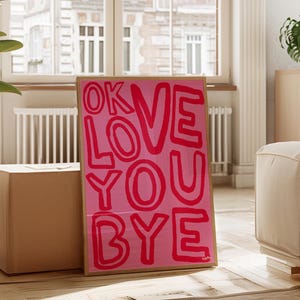 K&ouml;nnte beinhalten: Ein gerahmter rosa Kunstdruck mit den Worten "OK LOVE YOU BYE" in fetten roten Buchstaben. Das Kunstwerk wird auf einem Holzboden ausgestellt, der an einer Kiste lehnt, mit einem Fenster und einem cremefarbenen Sofa im Hintergrund.