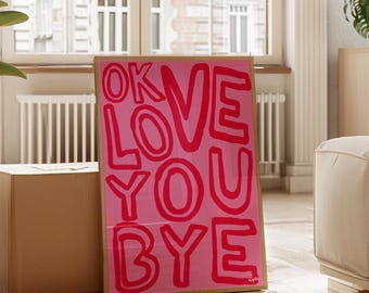 May include: A framed pink art print with the words "OK LOVE YOU BYE" in bold red letters. The artwork is displayed on a wooden floor, leaning against a box, with a window and a cream-colored sofa in the background.