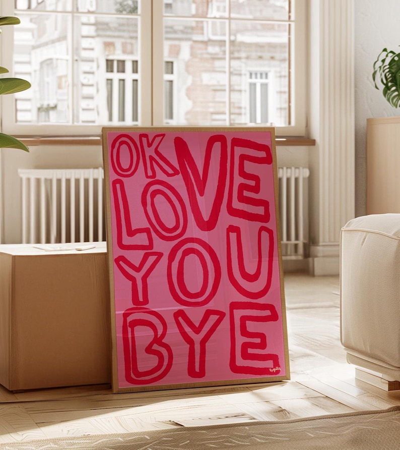 K&ouml;nnte beinhalten: Ein gerahmter rosa Kunstdruck mit den Worten "OK LOVE YOU BYE" in fetten roten Buchstaben. Das Kunstwerk wird auf einem Holzboden ausgestellt, der an einer Kiste lehnt, mit einem Fenster und einem cremefarbenen Sofa im Hintergrund.