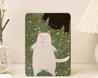 May include: A rectangular art print featuring two cats in a whimsical scene. A white cat stands upright in a field of green grass and colorful flowers. Above, a black cat hangs upside down. The artwork has a playful, cartoonish style.