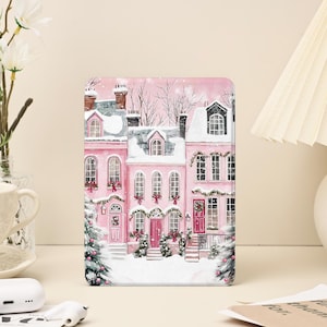 以下が含まれることがあります： ピンク色の長方形のケース。水彩画で雪に覆われた家々が描かれています。家には赤いドア、緑のリース、雪に覆われた屋根があります。ケースはクリスマスツリーとお祝いの装飾で飾られています。