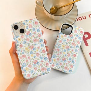 May include: Two white phone cases with a floral pattern featuring pink, blue, and yellow flowers. The cases are held in a person's hand. A teacup and saucer with a gold spoon are in the background.