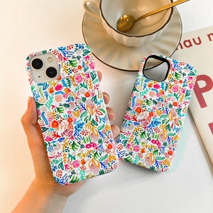 Może przedstawiać: Dwa białe etui na iPhone'a z kolorowym kwiatowym wzorem. Projekt przedstawia r&oacute;żnorodne kwiaty i liście w odcieniach r&oacute;żu, niebieskiego, pomarańczowego i zielonego. Etui są pokazane na białej powierzchni.