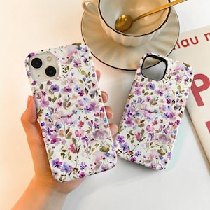 May include: Two floral phone cases with a white background. The cases feature a watercolor design of purple, pink, and blue flowers with green leaves. One case is held in a hand, and a teacup and saucer are in the background.