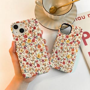 Op de afbeelding: Twee telefoonhoesjes met een bloemenpatroon. De hoesjes zijn wit met rode, gele en oranje bloemen. De hoesjes zijn gemaakt van hard plastic.