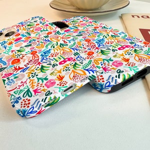 Może przedstawiać: Białe etui na telefon z kolorowym wzorem kwiatowym. Wz&oacute;r przedstawia r&oacute;żnorodne kwiaty i liście w odcieniach r&oacute;żu, pomarańczy, błękitu i zieleni. Etui mają czarne krawędzie i wycięcia na aparat i przyciski.