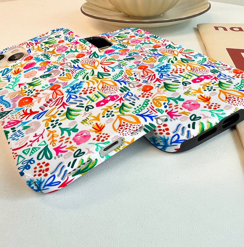 Może przedstawiać: Białe etui na telefon z kolorowym wzorem kwiatowym. Wz&oacute;r przedstawia r&oacute;żnorodne kwiaty i liście w odcieniach r&oacute;żu, pomarańczy, błękitu i zieleni. Etui mają czarne krawędzie i wycięcia na aparat i przyciski.