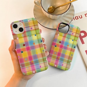 May include: Two colorful phone cases with a plaid pattern in pink, yellow, green, and blue. The cases are decorated with silver and colorful star shapes. One case is held in a hand, and the other is lying on a white surface.