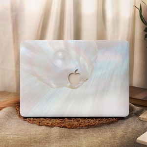 May include: A laptop skin featuring a pearlescent, iridescent shell pattern with a soft white and light blue color palette. The design displays a smooth, glossy finish with a circular pearl graphic positioned above the central Apple logo cutout.