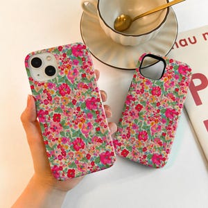 May include: Two floral patterned phone cases with a vibrant mix of pink, red, yellow, and green flowers on a white background. The cases are held in a person's hand. A teacup and saucer are in the background.