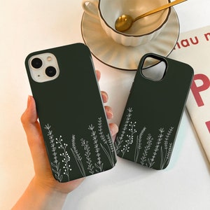 May include: Two dark green phone cases with a white floral design. The cases are shown side by side, one held in a hand. A cup of coffee and a book are in the background.