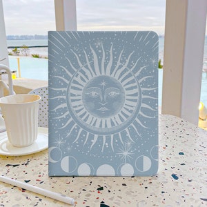 May include: A light blue notebook with a white sun and moon design. The sun has a face and the moon is in various phases. The background is a light blue with white stars.