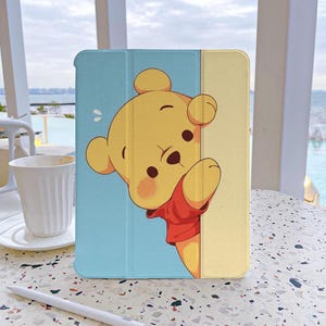 May include: A blue and yellow tablet case with a cartoon image of Winnie the Pooh peeking around a corner. The case has a fold-over flap for protection.