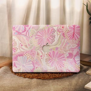 Puede incluir: Una funda rígida para portátil con un vibrante estampado floral en tonos rosa, crema y beige claro. El diseño incluye líneas intrincadas, texturas punteadas y motivos florales estilizados, con un recorte central para el logotipo de Apple.