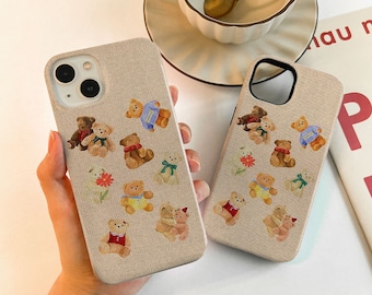 Puede incluir: Dos fundas de teléfono beige con un diseño de osito de peluche marrón y blanco. Las fundas tienen un acabado de tela texturizada y un recorte para la cámara.
