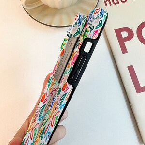 Może przedstawiać: Białe etui na telefon z kolorowym kwiatowym wzorem z r&oacute;żowymi, pomarańczowymi, niebieskimi i zielonymi akcentami. Etui ma czarną obw&oacute;dkę i srebrny przycisk. Projekt to żywy, artystyczny wyświetlacz.