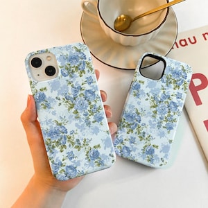 Puede incluir: Dos fundas de teléfono con un estampado floral azul claro con rosas azules y hojas verdes. Las fundas están en la mano de una persona. Una taza y un platillo blancos con una cuchara dorada están en el fondo.