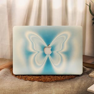Op de afbeelding: Een laptophoes met een zacht gefocuste, airbrush-stijl vlindergrafiek in tinten lichtblauw en wit. Het ontwerp is gecentreerd rond de uitsparing voor het Apple-logo, dat zichtbaar blijft als een zilverkleurig metalen icoon in het midden.