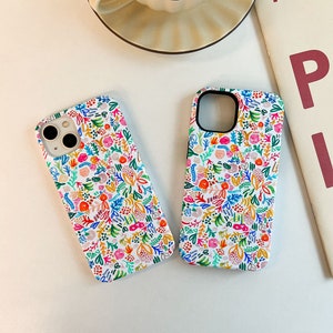 Może przedstawiać: Dwa białe etui na telefon z kolorowym kwiatowym wzorem. Wz&oacute;r zawiera r&oacute;żowe, pomarańczowe, niebieskie i zielone kwiaty i liście. Etui mają czarne wycięcia na aparat i białą obw&oacute;dkę.