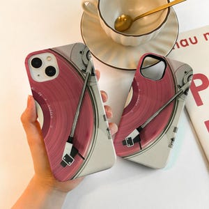 May include: Two phone cases featuring a pink vinyl record design with a silver tone turntable arm. The cases have a white and gray border. The text "All Of A Sudden" is visible on the record. A cup and saucer are in the background.