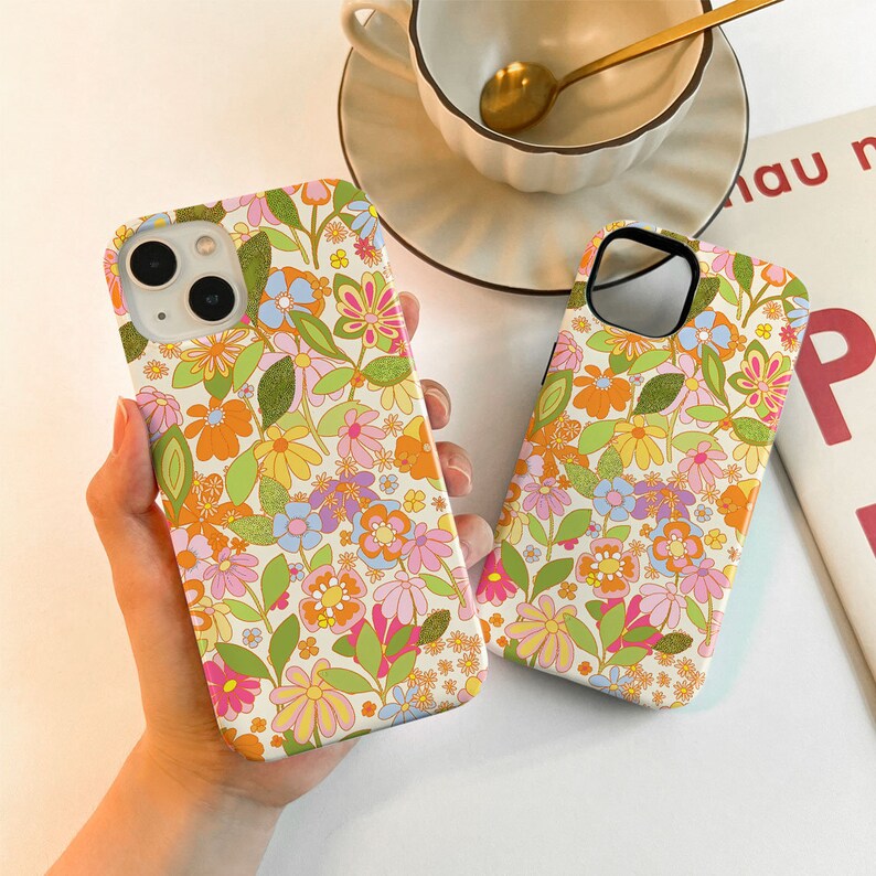 May include: Two phone cases featuring a floral design. The cases are white with a variety of pink, orange, yellow, and green flowers. The cases are designed to fit a smartphone.